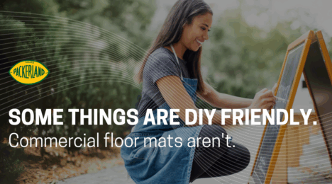 A woman with brown, shoulder length hair, wearing jeans and a grey t-shirt kneels on the ground in front of a chalkboard. She is writing the specials of the day on the chalkboard. Over the image, text reads, "Some things are DIY friendly. Your commercial floor mats aren't."