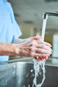 A person is washing their hands in a stainless steel sink, seemingly in a hospital environment as they are wearing light blue scrubs. Their hands are the focus of the photo, so no other identifying information is shown.