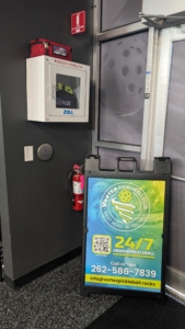 An AED is shown hanging on a dark wall to the left of a banner for a Pickleball club sign. Also in the frame is a fire extinguisher.