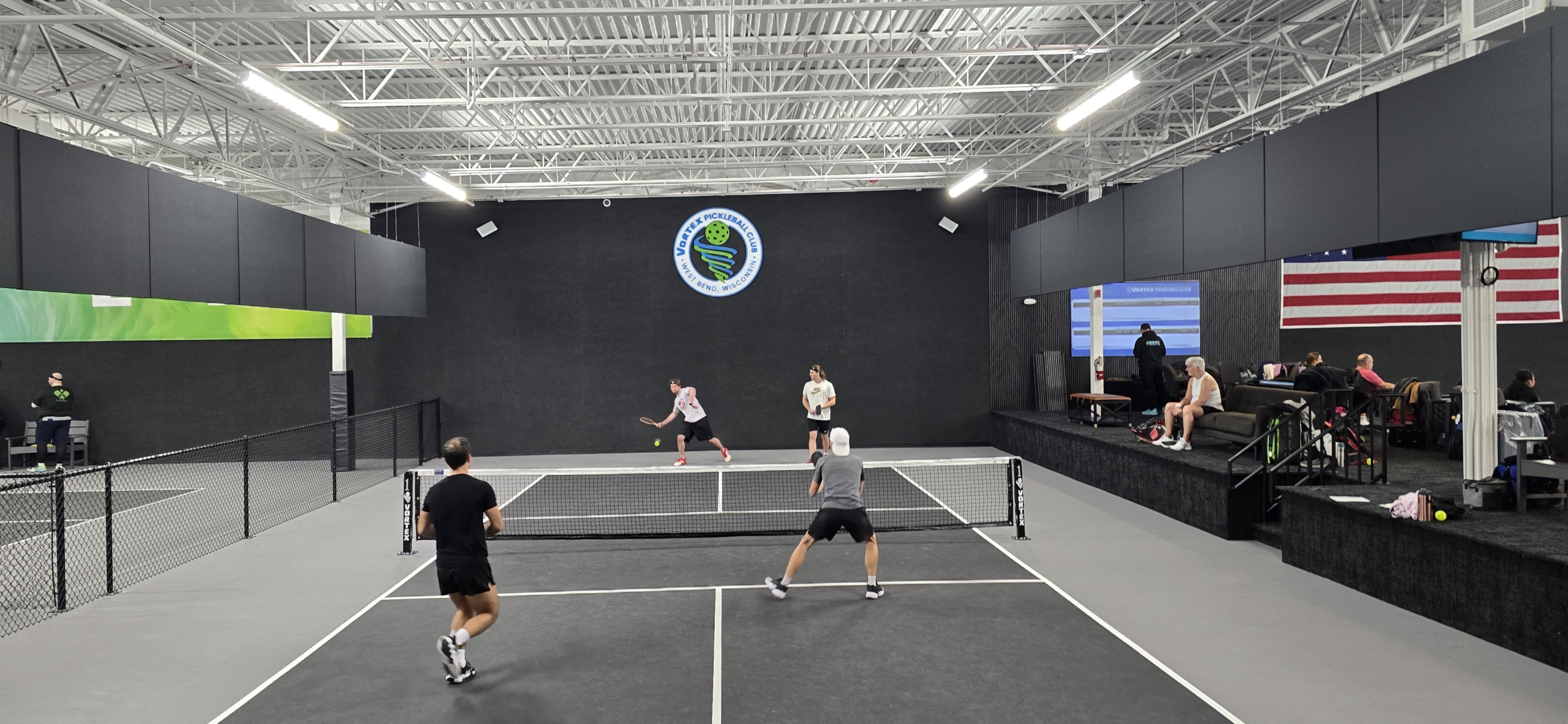 20250222_103530 An indoor pickleball court is shown surrounded by dark grey walls and bright, white ceiling. On the court, three players are actively involved in a game, running, volleying, and scanning the court for their next move. On the wall is Vortex Pickleball's green and blue logo.
