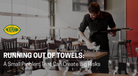 Packerland's blog header image contains the Heading of the blog, Running Out of Towels; a Small Problem That Can Create Big Risks. Behind the title, a restaurant employee is using a towel from a commercial towel service to wipe down empty tables
