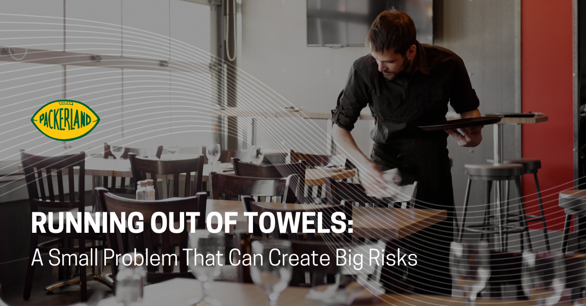 Packerland's blog header image contains the Heading of the blog, Running Out of Towels; a Small Problem That Can Create Big Risks. Behind the title, a restaurant employee is using a towel from a commercial towel service to wipe down empty tables