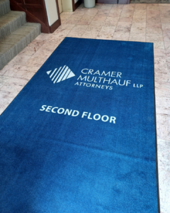 Blue custom logo mat for Cramer Multhauf LLP Attorneys directing visitors to the second floor, placed in a tiled lobby near a carpeted staircase.