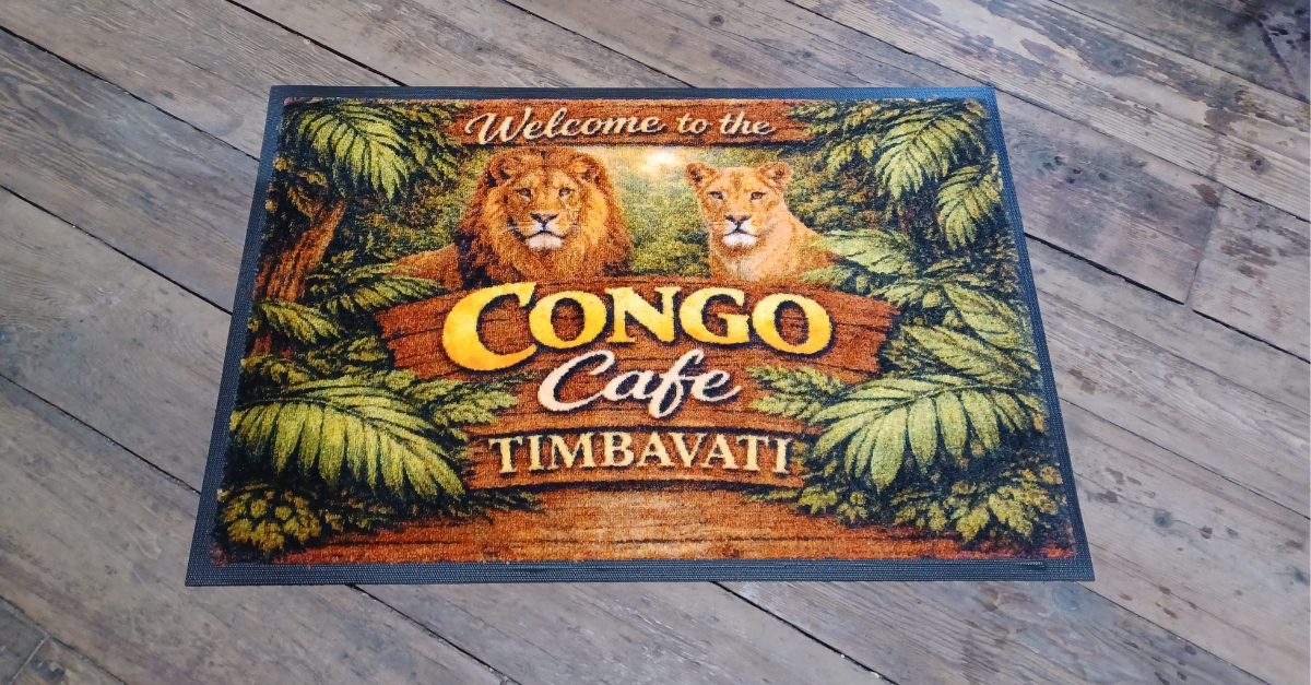 Custom logo mat welcoming guests to the Congo Cafe at Timbavati, featuring a lion and lioness surrounded by jungle foliage, placed on a rustic wood plank floor.