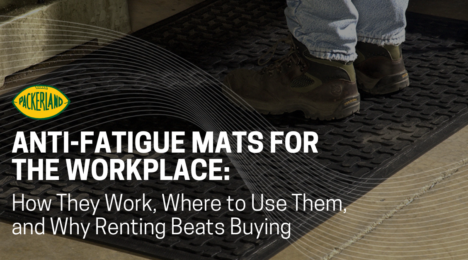 A man's feet are shown wearing steel toed boots stands on a black commercial anti-fatigue floor mat.