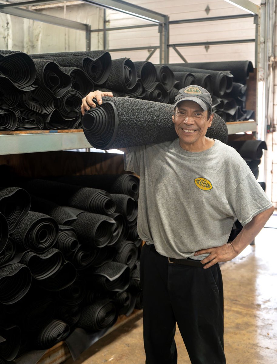 A smiling Packerland team member carries a rolled entrance mat past racks of mats.